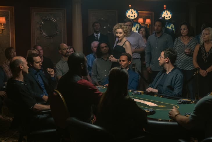 People gathered around a poker table in a dimly lit room, with a woman standing and observing the game.