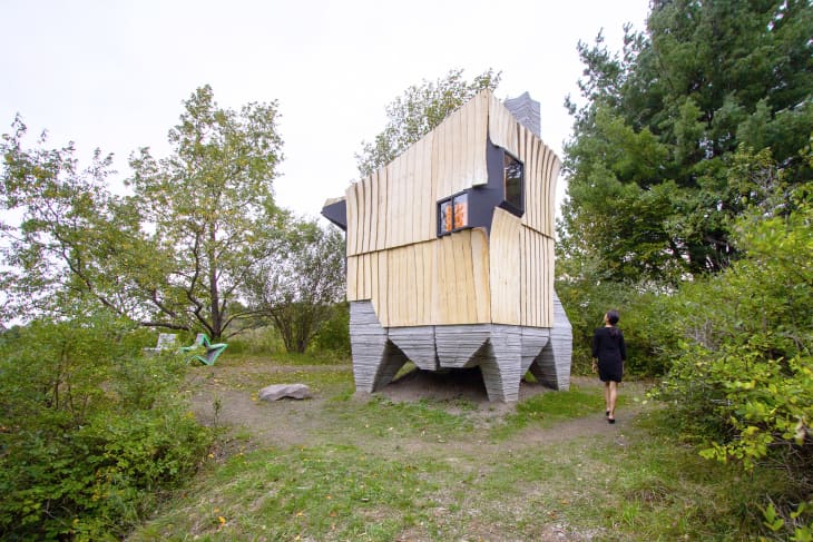 Unique wooden cabin with angular design in a forest setting, person walking towards it.