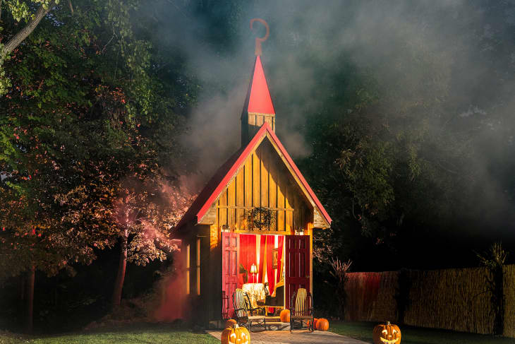 Small wooden chapel with red roof, surrounded by trees and fog, lit pumpkins on porch.
