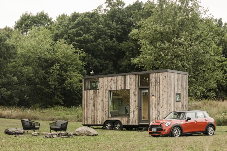 Tiny house with wooden exterior and large window, parked on grass beside an orange car, surrounded by trees and outdoor chairs.