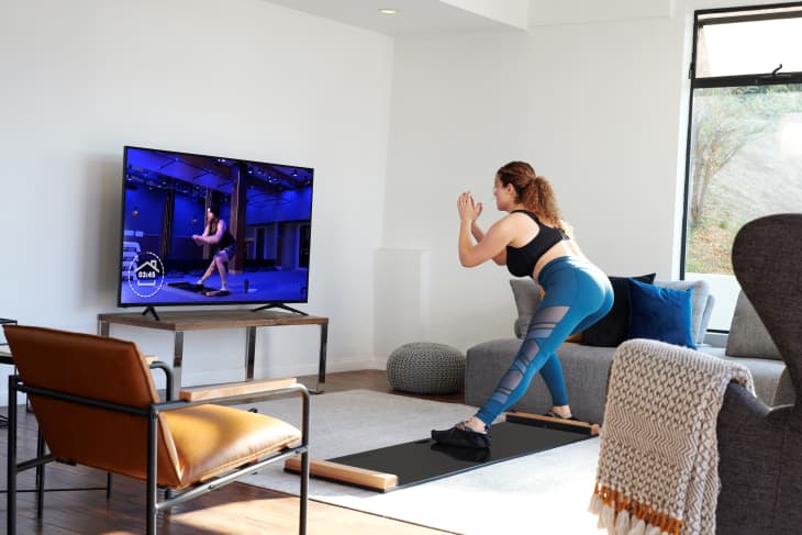Woman exercising in living room, following a workout video on TV, with modern furniture and large window.