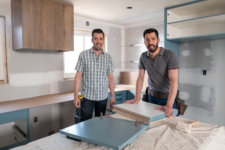 Two men in a partially renovated kitchen with blue cabinets and wooden shelves, holding a cabinet door.