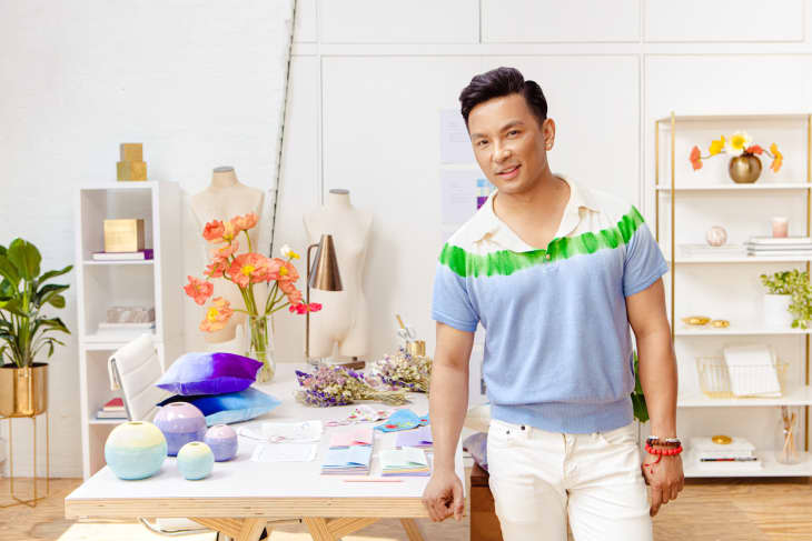 Man in a blue and green shirt standing by a table with colorful decor items, flowers, and mannequins in a bright studio.