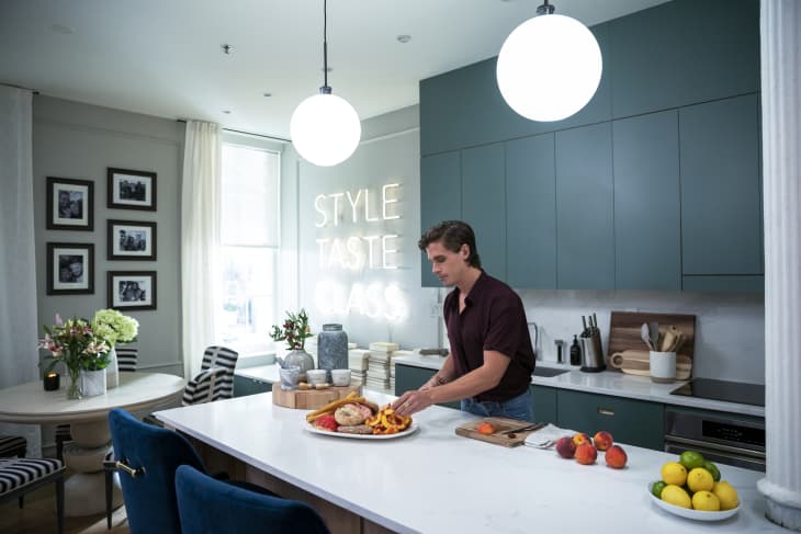 Man preparing food in a modern kitchen with teal cabinets, white countertops, and a neon sign reading "STYLE TASTE CLASS."