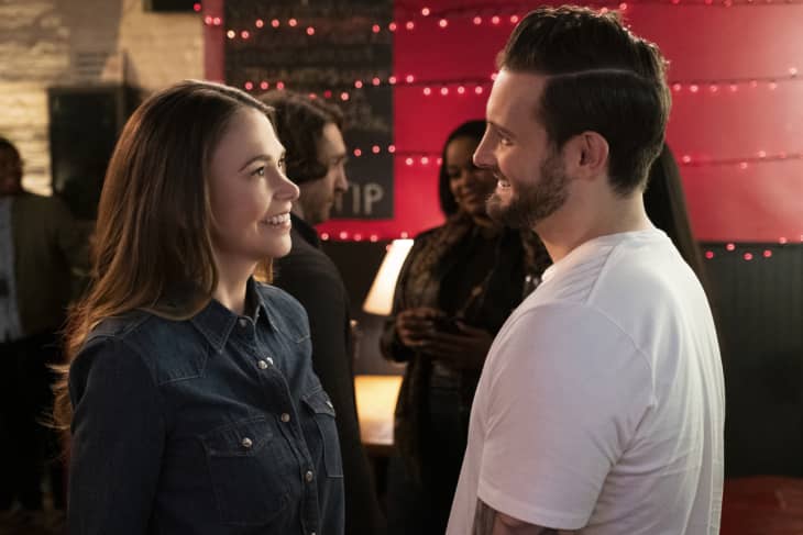 A woman in a denim shirt and a man in a white t-shirt smiling at each other in a dimly lit room with red string lights.