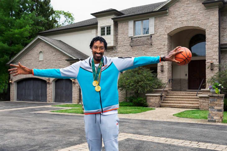 Man in a blue tracksuit holding a basketball, standing in front of a large stone house with medals around his neck.