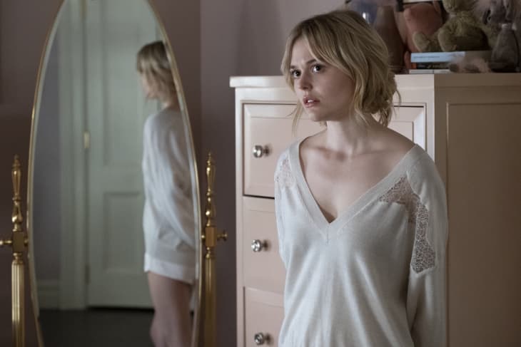 Woman in a white sweater standing by a dresser, with her reflection visible in a gold-framed mirror.