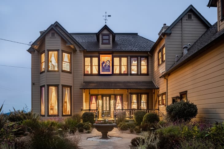 Victorian-style house with large bay windows, lit interior, and a central garden fountain at dusk.