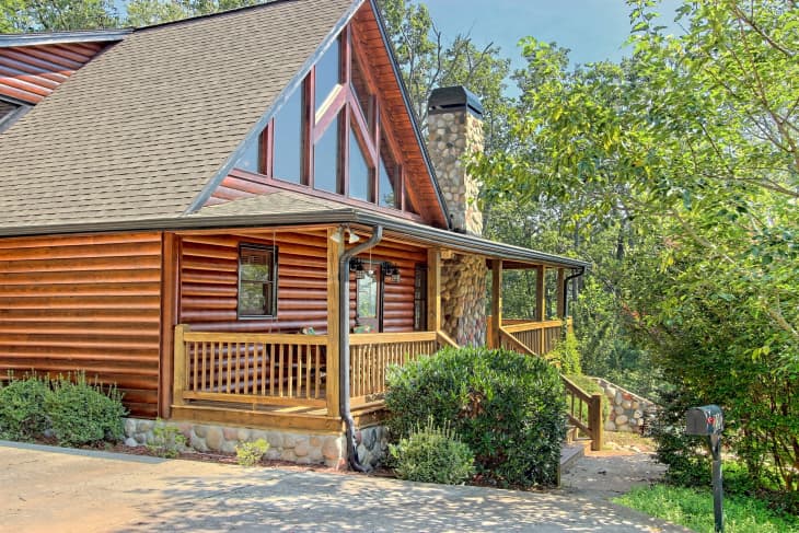Log cabin with large triangular windows, stone chimney, and wooden porch surrounded by trees and greenery.