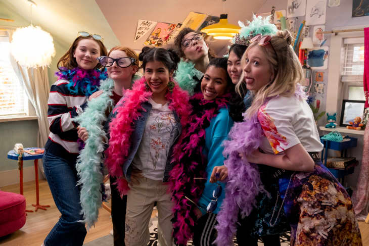 Group of six friends in a colorful room, wearing feather boas and playful accessories, posing for a fun photo.
