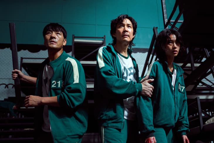 Three people in green tracksuits with numbers, standing under metal stairs, looking tense.