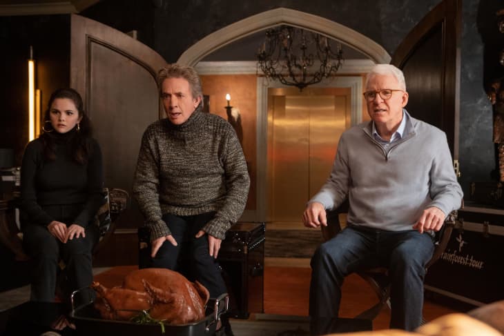 Three people sitting in a dimly lit room with a roasted turkey on a table, chandelier overhead.
