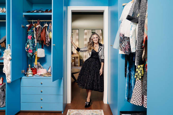 Woman in a black dress standing in a blue walk-in closet with open wardrobe doors displaying clothes and accessories.