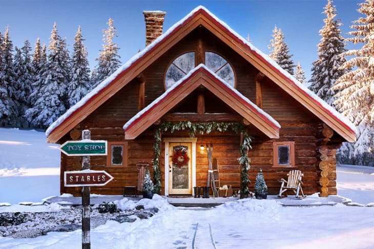Log cabin in snowy forest with wreath on door, skis, and wooden chairs. Signpost points to toy shop and stables.