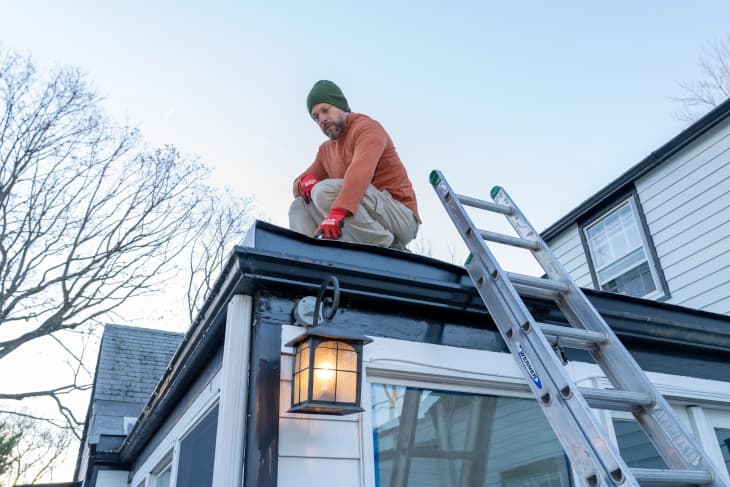 Man in orange sweater and green beanie sitting on roof edge, next to a ladder and outdoor lantern, with bare trees in background.