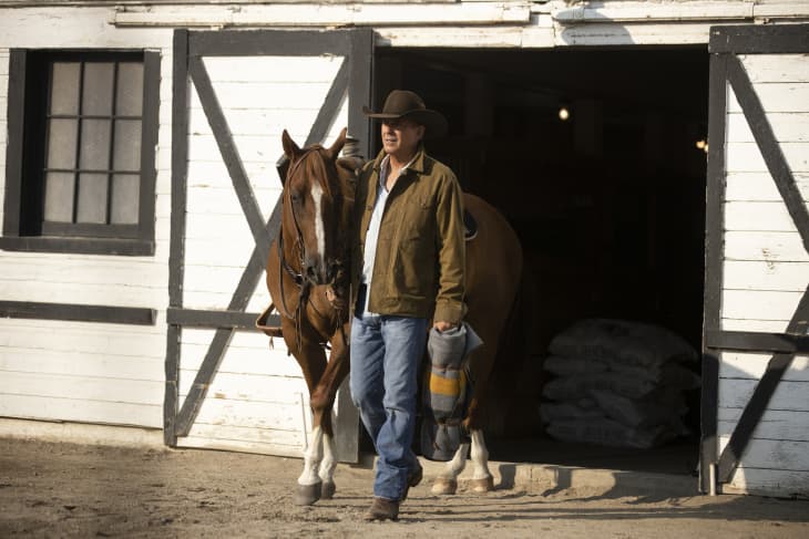 Man in cowboy hat leading a horse out of a white barn with black trim.