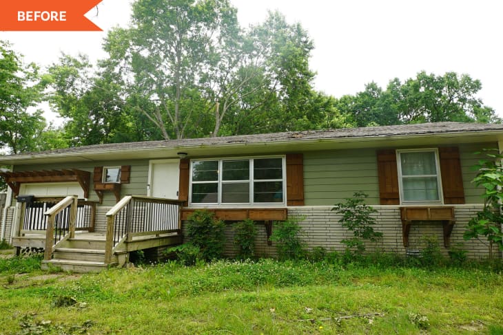 Single-story house with green siding, wooden shutters, front porch, and overgrown yard, surrounded by trees.