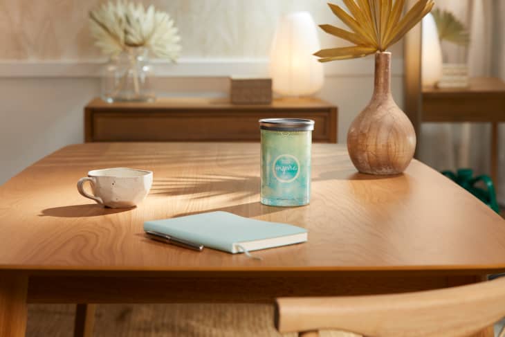 Wooden table with a candle, ceramic mug, notebook, and decorative vase in a softly lit room.