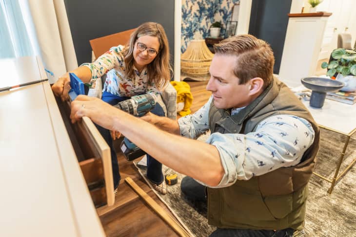 Two people assembling furniture with a drill in a living room, featuring a floral wallpaper and modern decor.