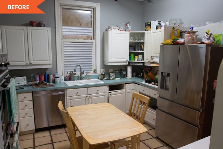 Cluttered kitchen with white cabinets, stainless steel fridge, wooden table, and various items on countertops.