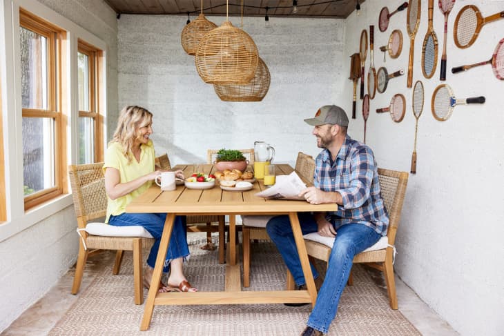 Couple enjoying breakfast at a wooden table in a sunroom with wicker chairs and vintage tennis rackets on the wall.