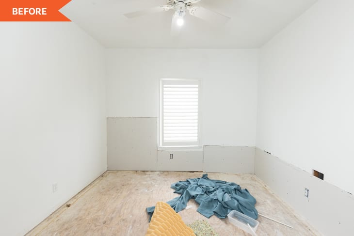 Empty room under renovation with white walls, plywood floor, ceiling fan, and window with shutters.