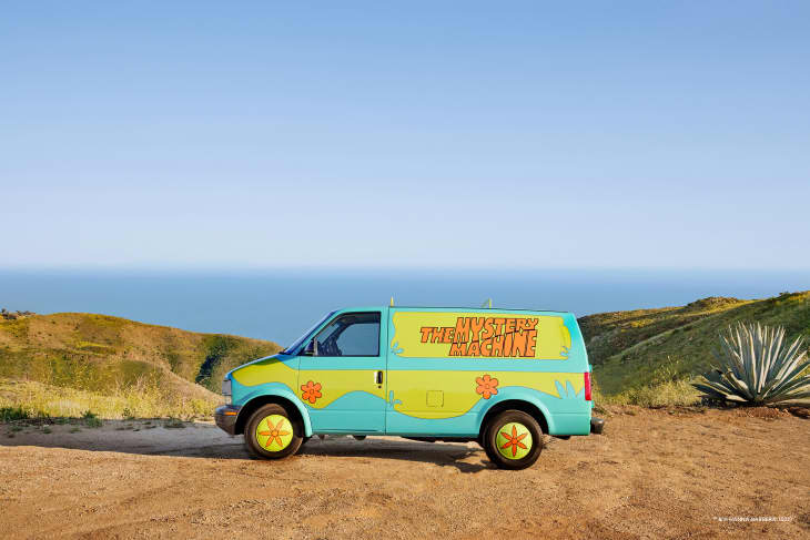 Colorful van labeled "Mystery Machine" parked on a dirt path with ocean and hills in the background.