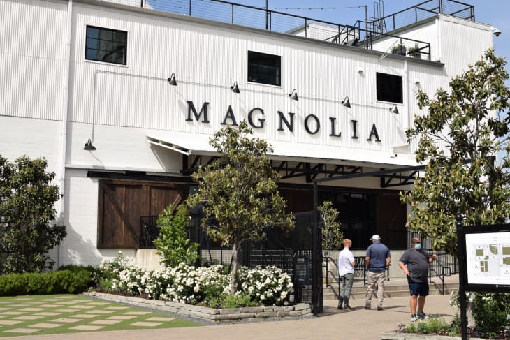 Exterior of Magnolia building with large sign, surrounded by trees and flowers, and three people walking towards the entrance.
