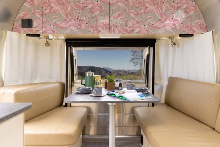 Camper interior with beige seating, flamingo wallpaper, and a table set with dishes and a green thermos, overlooking a scenic view.