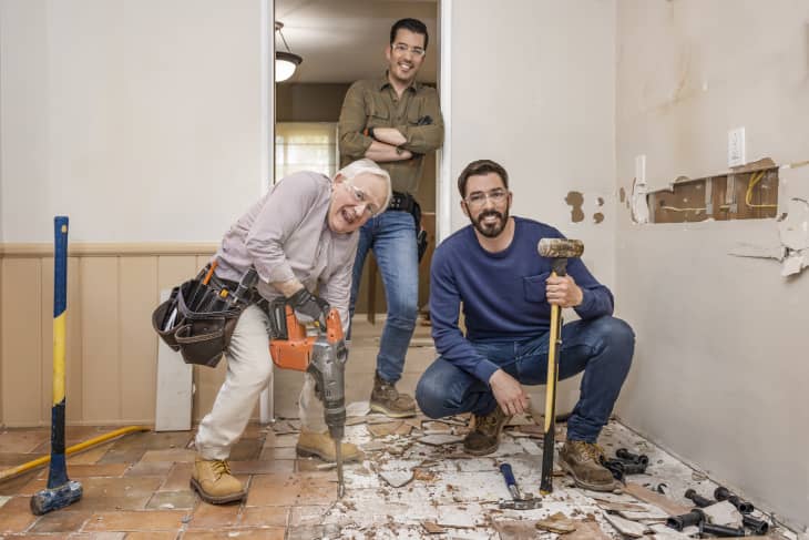 Three men in a partially demolished room, one using a jackhammer, another holding a sledgehammer, and debris on the floor.