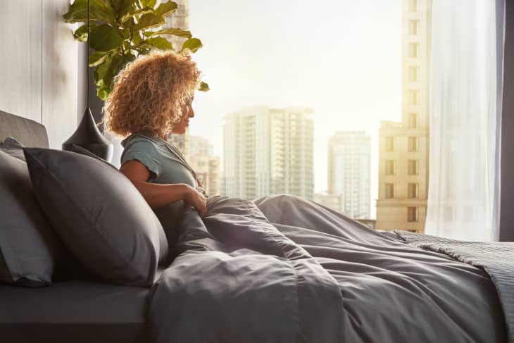 Woman with curly hair sitting on a bed in a modern bedroom, cityscape visible through large window.