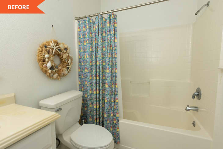 Small bathroom with floral shower curtain, white bathtub, toilet, and seashell wreath on the wall.