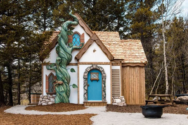 Fairy-tale cottage with a blue door, beanstalk decor, and wooden shingles, surrounded by trees and a picnic area.