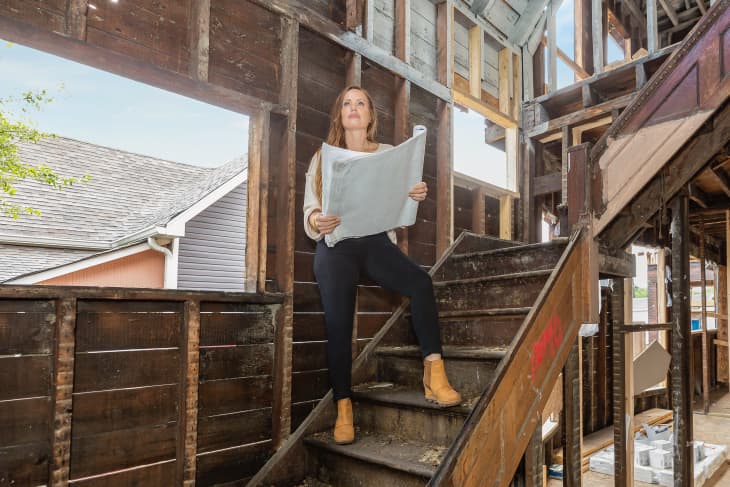 Woman holding blueprints on wooden stairs in a house under renovation.