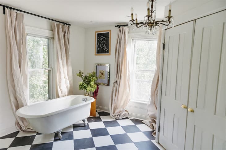 Freestanding bathtub in a bright bathroom with checkered floor, large windows, beige curtains, and a chandelier.