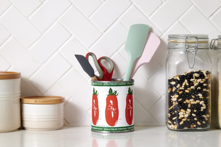 tomato can holding kitchen tool items on a kitchen counter with 2 white containers with wooden lids and a glass of back and white peas next to it. and a tiled backdrop