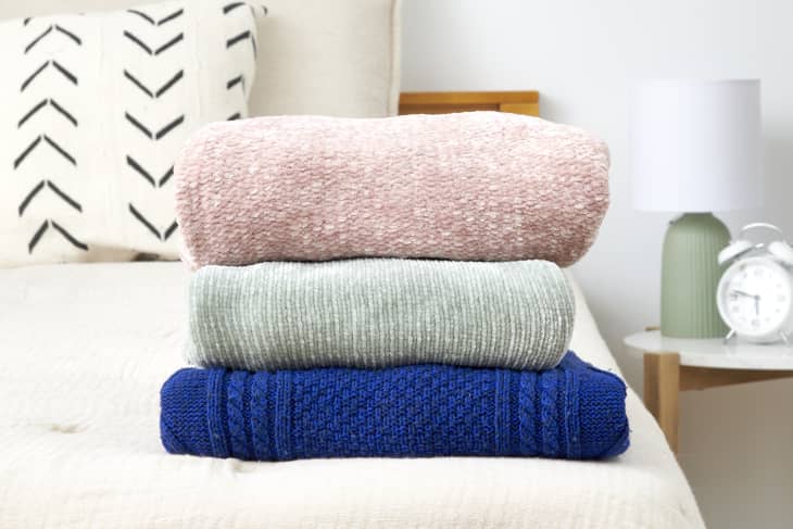 Close up view of a stack of three sweaters on a beige bed, with a green lamp and white alarm clock on a wood and marble table in the background.