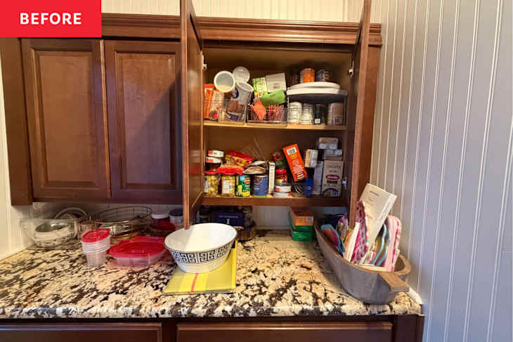 Cluttered kitchen cabinet with various canned goods, containers, and a bowl on a granite countertop.