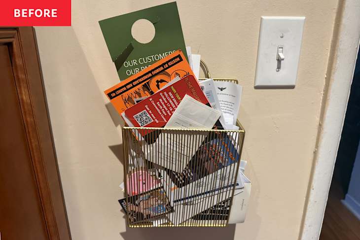 A gold wire basket filled with various flyers and papers mounted on a wall next to a light switch.