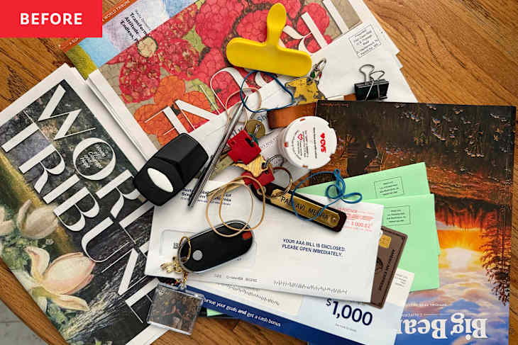 A cluttered wooden table with magazines, keys, a pill bottle, paper clips, and various envelopes.