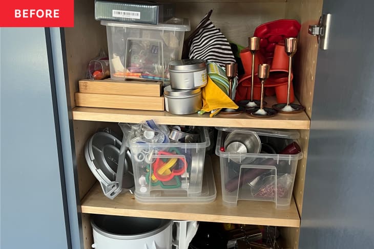 Cluttered cabinet with plastic bins, candles, baking tools, and kitchen items on wooden shelves.