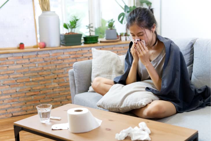 Sick woman sneezing at home on the sofa with a cold, she is covering with a blanket and blowing her nose