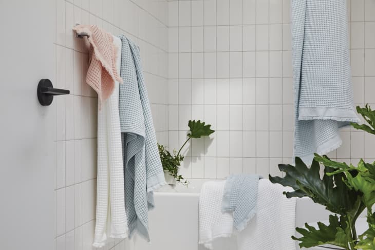 Bathroom with white tiles, pastel towels hanging, and green plants around a bathtub.
