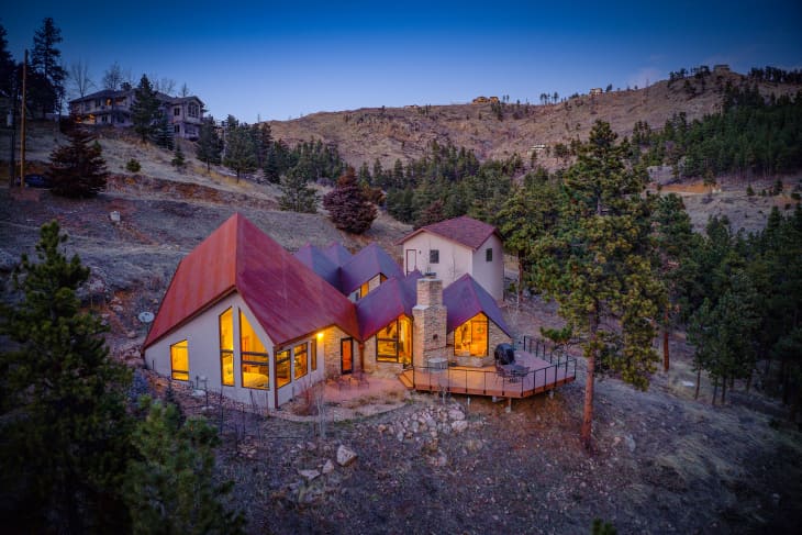 A-frame house with red roof, large windows, and outdoor deck nestled in a wooded hillside at dusk.