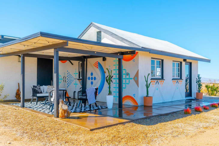 Desert house with colorful geometric mural, covered patio, black and white furniture, and potted plants.
