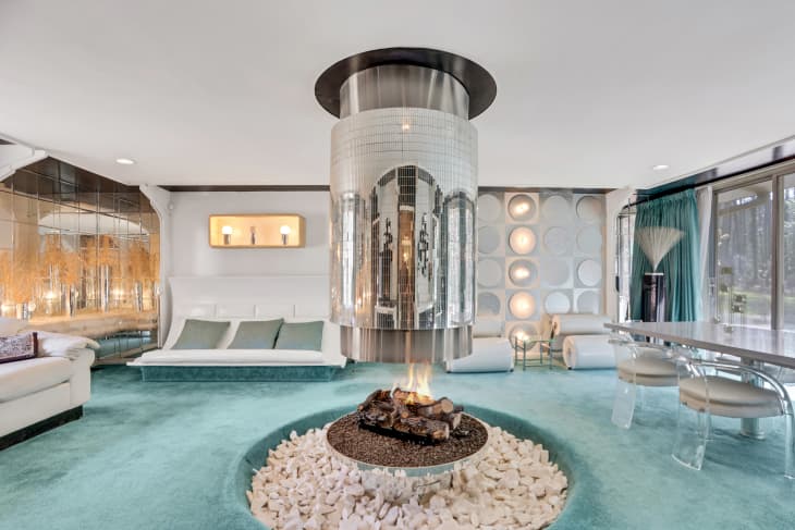 Retro living room with a central mirrored fireplace, turquoise carpet, white seating, and circular wall decor.