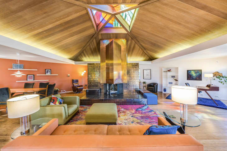 Mid-century modern living room with orange sofa, colorful rug, central fireplace, and wooden ceiling.