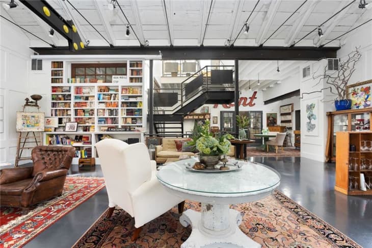 Loft-style living room with bookshelves, leather armchair, white armchair, round table, and staircase leading to upper level.