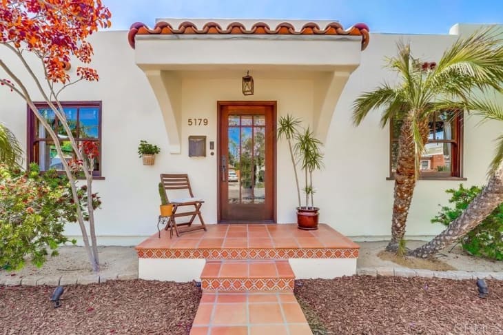 Spanish-style house entrance with terracotta tiles, wooden chair, potted plants, and arched doorway.
