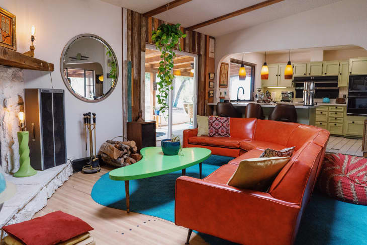 Retro living room with orange sectional sofa, green coffee table, round mirror, and open kitchen with pendant lights.